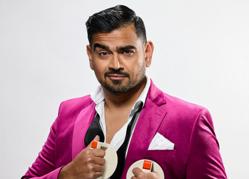 Dilruk Jayasinha in a pink suede suit, an undone tie around his neck, holding defibrillator paddles against his chest. and 