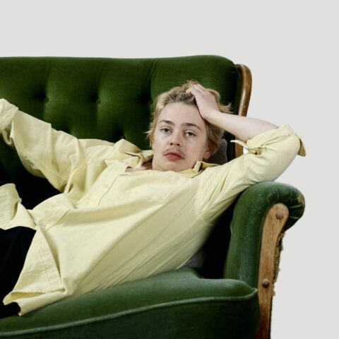 A white person is lying on a green couch staring directly at the camera. They have short dirty blonde hair with dark eyebrows and moustache. They are wearing a light yellow dress shirt. 