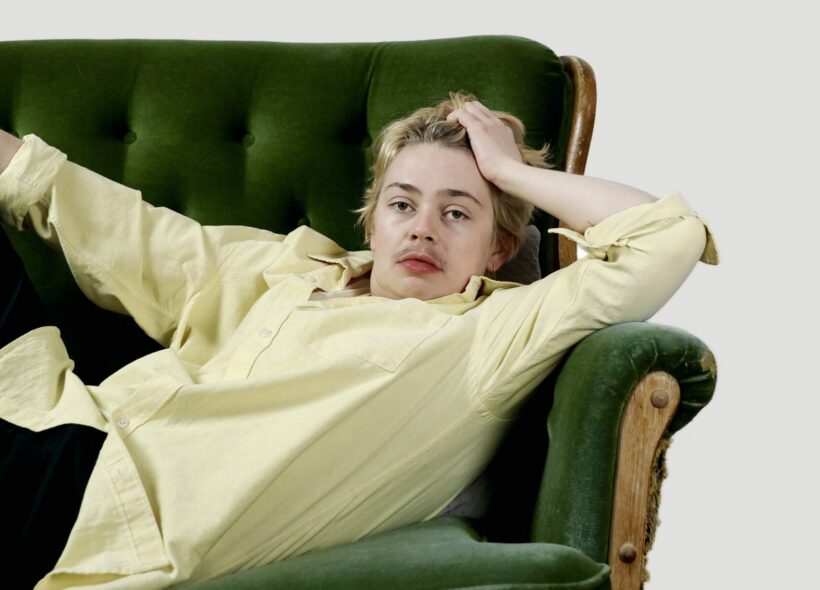 A white person is lying on a green couch staring directly at the camera. They have short dirty blonde hair with dark eyebrows and moustache. They are wearing a light yellow dress shirt. 