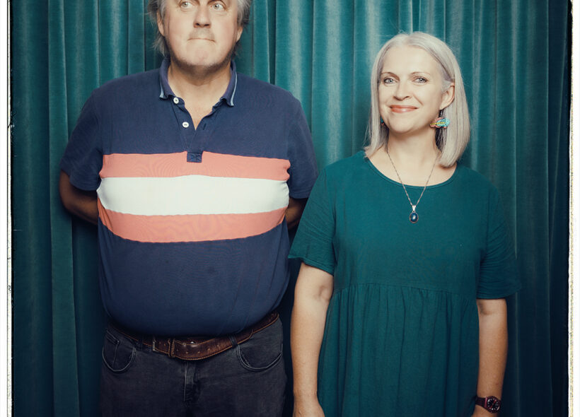 An adult brother and sister stand beside each other in front of a green curtain.