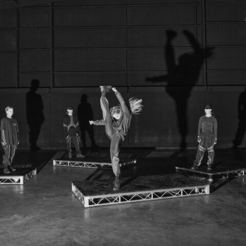 Four dancers dressed in black streetwear stand on low, rectangular staging decks against the backdrop of an expansive black warehouse. The dancer at the centre of the image is doing a high kick while her hair flicks behind her.