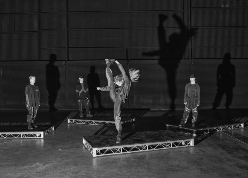 Four dancers dressed in black streetwear stand on low, rectangular staging decks against the backdrop of an expansive black warehouse. The dancer at the centre of the image is doing a high kick while her hair flicks behind her.
