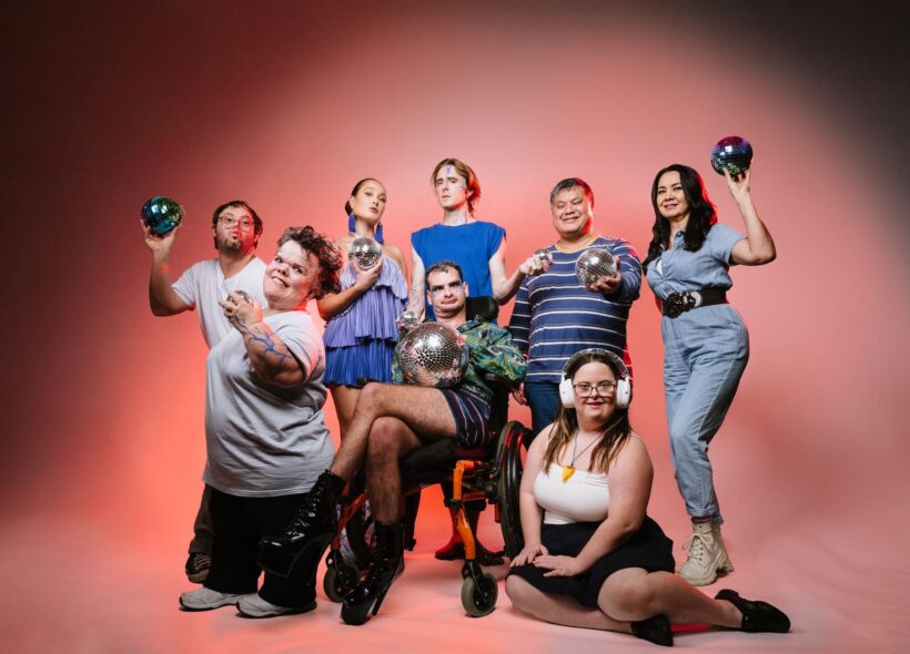 A group of artists from UAF posing together infront of an orange wall. They are in various poses with different size silver disco balls in their hands. 