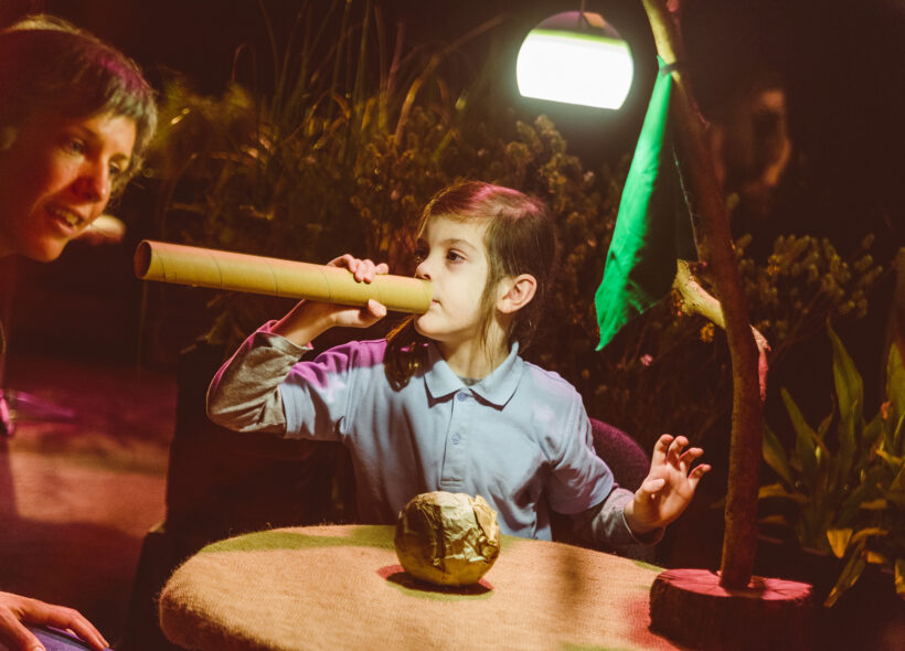 A When the World Turns production photo. A child holds up a cardboard tube, using it to make sounds. An adult leans towards the tube, listening. They sit at a small table in a darkened space, surrounded by plants. They are illuminated by a lamp on the table. Photographer: Theresa Harrison
