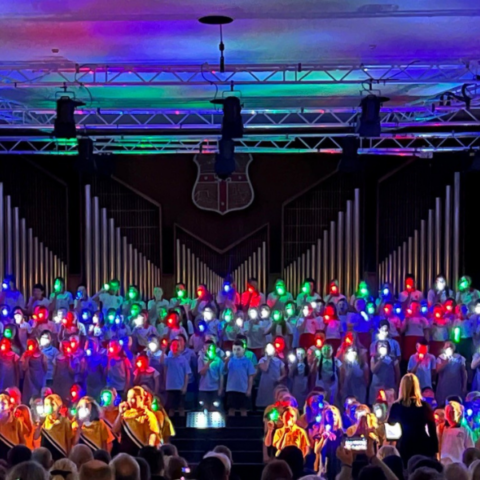 A choir performance in a dimly lit auditorium features singers holding colourful lanterns of red and green. They stand in front of a large organ backdrop. A conductor, with arms raised, is directing the choir. The audience in the foreground is barely visible.