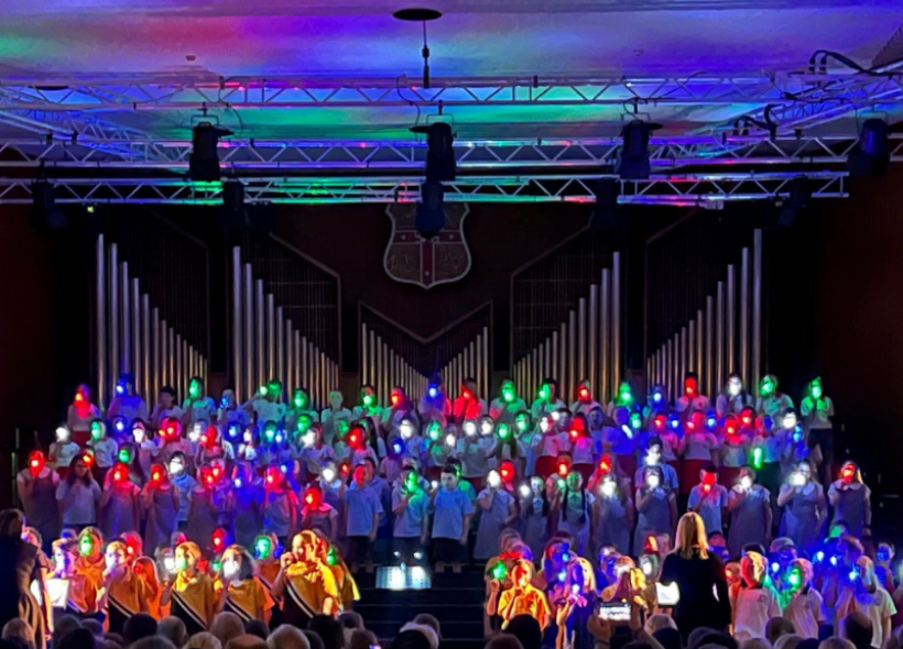 A choir performance in a dimly lit auditorium features singers holding colourful lanterns of red and green. They stand in front of a large organ backdrop. A conductor, with arms raised, is directing the choir. The audience in the foreground is barely visible.