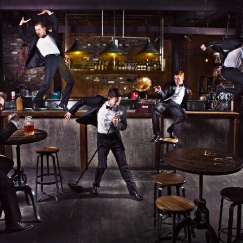 Tap dance troupe scattered around a dimly lit bar wearing tuxedos.