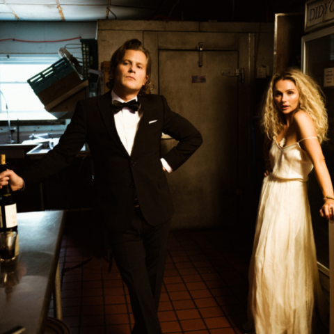Two people in an industrial kitchen wearing formal tuxedo and dress wear.