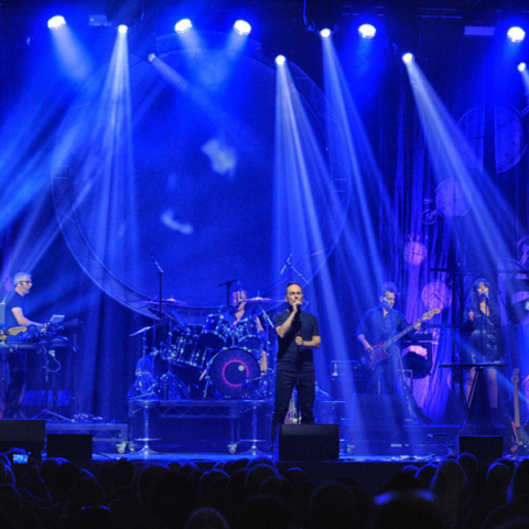 Seven band members on a blue lit stage performing to a crowd of people.