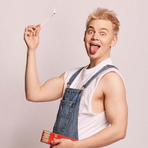 Joel Creasy in denim overalls and a white t-shirt holding a lit match with his tongue sticking out.