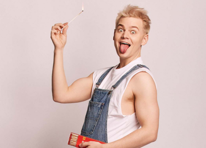Joel Creasy in denim overalls and a white t-shirt holding a lit match with his tongue sticking out.