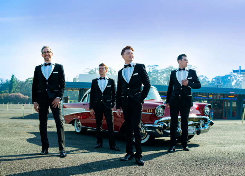 The Vallies dressed in tuxedos in a diner car park with a vintage red sports car.
