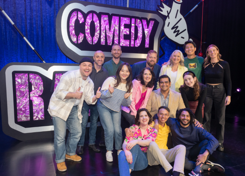 RAW Comedy contestants huddled on stage together in front of RAW Comedy purple signage