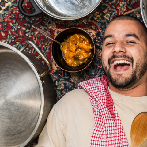 A person with a cheerful expression lies on a patterned rug next to a pot and a bowl of food. The bowl contains a dish with yellow sauce and chunks, and a spoon is placed inside. The scene exudes a sense of joy and culinary enjoyment.
