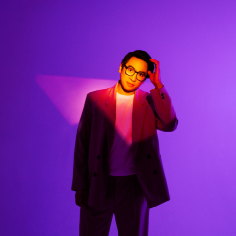 A person in a stylish suit and glasses stands against a vibrant purple background, with dramatic lighting casting shadows and highlights.