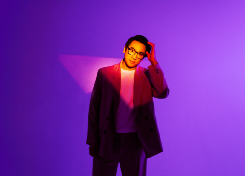A person in a stylish suit and glasses stands against a vibrant purple background, with dramatic lighting casting shadows and highlights.