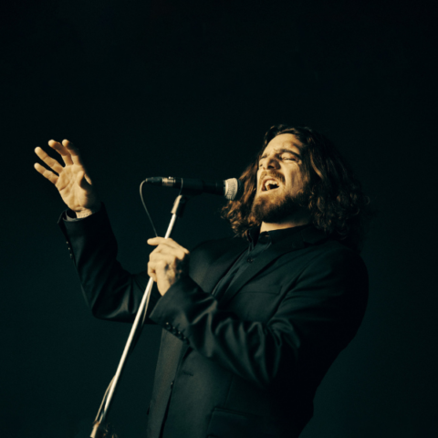 Man with long wavy hair in a black long sleeve shirt singing into a microphone on a stand.