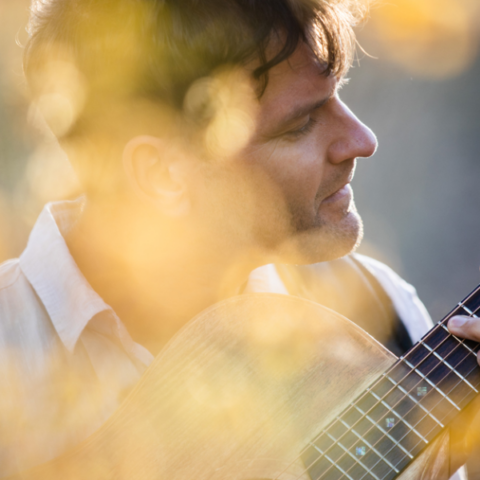 A person with short hair is playing an acoustic guitar among blurred golden-yellow foliage. The soft sunlight creates a warm and serene atmosphere. The person's eyes are closed or looking downward, suggesting a focus on the music.