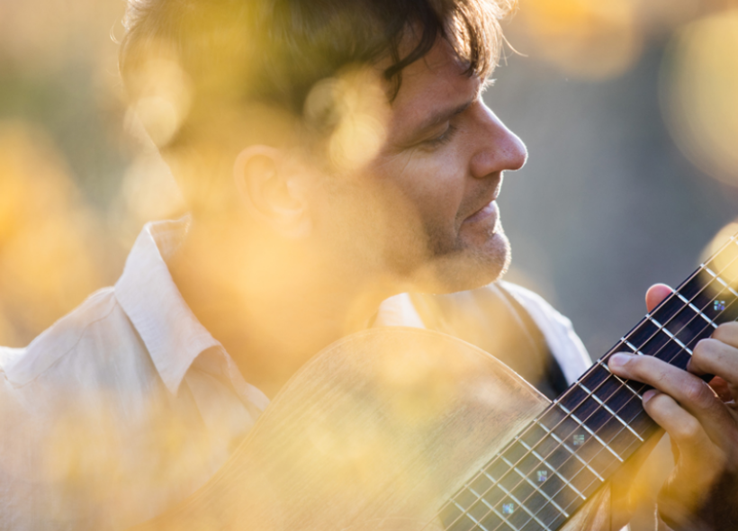 A person with short hair is playing an acoustic guitar among blurred golden-yellow foliage. The soft sunlight creates a warm and serene atmosphere. The person's eyes are closed or looking downward, suggesting a focus on the music.