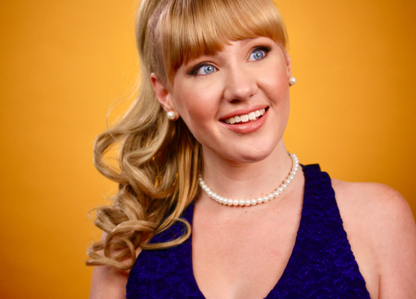 A smiling woman with blue eyes and blonde hair styled in loose curls. She is wearing a pearl necklace, pearl earrings, and a sleeveless blue dress, with an orange background behind her.