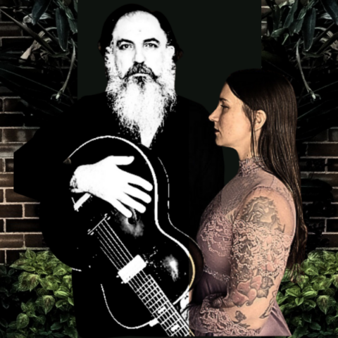 Bearded man with guitar and tattooed woman in lacy dress against a brick wall with lush plants.