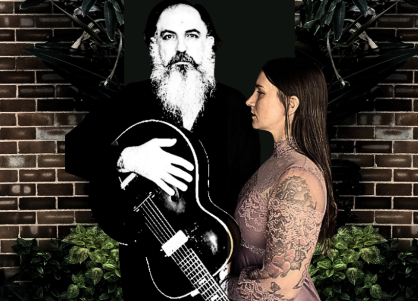 Bearded man with guitar and tattooed woman in lacy dress against a brick wall with lush plants.