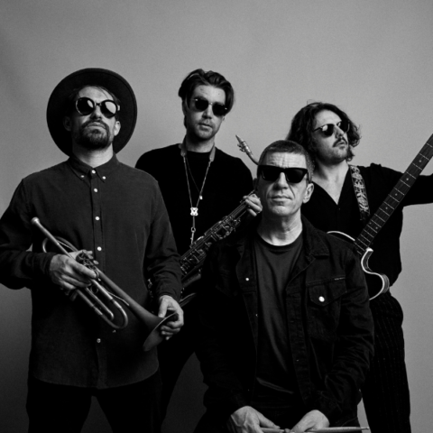 A black-and-white image of four men posing with musical instruments. The man on the left wears a hat and sunglasses, holding a trumpet. Next to him, a man in sunglasses holds a saxophone. A seated man and another standing man with a guitar also wear sunglasses.