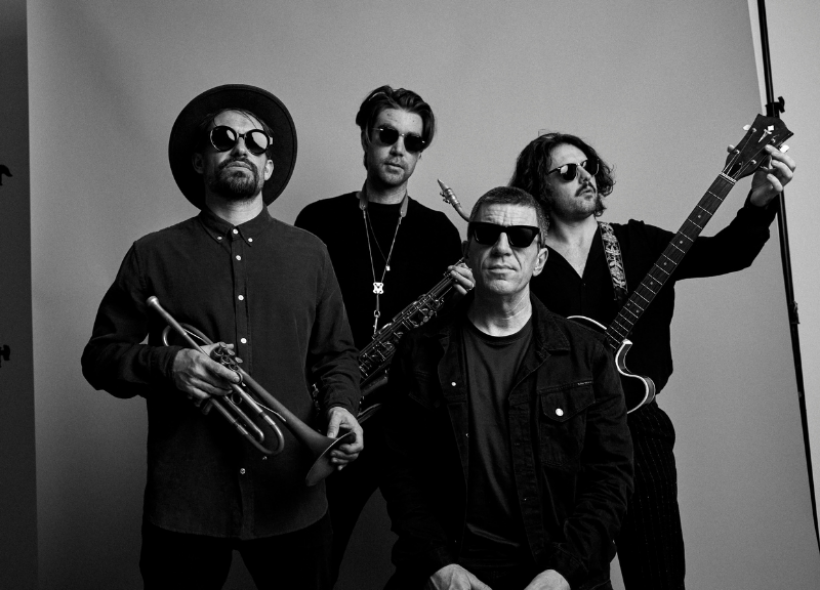 A black-and-white image of four men posing with musical instruments. The man on the left wears a hat and sunglasses, holding a trumpet. Next to him, a man in sunglasses holds a saxophone. A seated man and another standing man with a guitar also wear sunglasses.