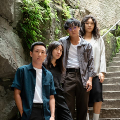 Four individuals stand together on a stone staircase with green moss growing on the walls. They all face the camera with neutral expressions, dressed in casual attire, with the person closest to the front wearing glasses. The scene appears outdoors in a natural setting.