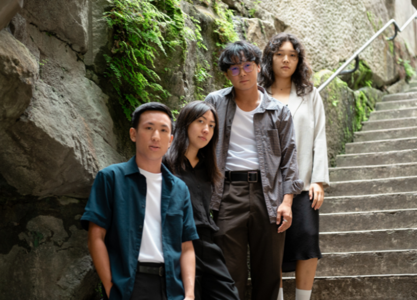 Four individuals stand together on a stone staircase with green moss growing on the walls. They all face the camera with neutral expressions, dressed in casual attire, with the person closest to the front wearing glasses. The scene appears outdoors in a natural setting.