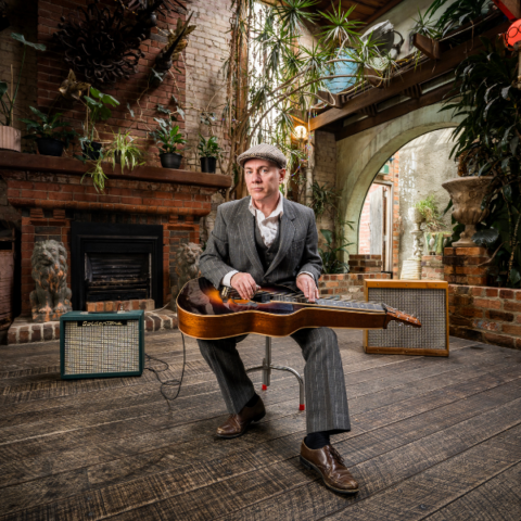 A person in a gray suit and flat cap sits on a stool in front of a fireplace, holding a vintage guitar. They are surrounded by potted plants and flanked by two guitar amplifiers on a wooden floor in a rustic indoor setting.