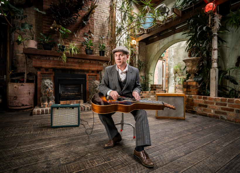 A person in a gray suit and flat cap sits on a stool in front of a fireplace, holding a vintage guitar. They are surrounded by potted plants and flanked by two guitar amplifiers on a wooden floor in a rustic indoor setting.