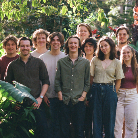 A group of ten people stands together in an outdoor garden surrounded by lush greenery. They are all smiling and dressed in casual clothing, creating a relaxed and friendly atmosphere. The background features various plants and trees, adding to the scenic setting.