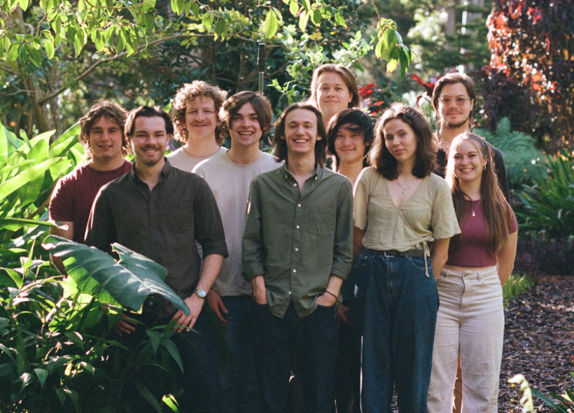 A group of ten people stands together in an outdoor garden surrounded by lush greenery. They are all smiling and dressed in casual clothing, creating a relaxed and friendly atmosphere. The background features various plants and trees, adding to the scenic setting.