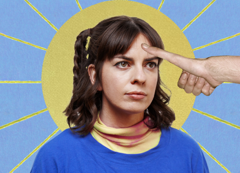 A woman with brown hair in pigtails looks serious while a hand extends from the right side of the image, with its index finger pointing at her forehead. The background features a circular yellow halo with sun rays radiating from it against a blue backdrop.