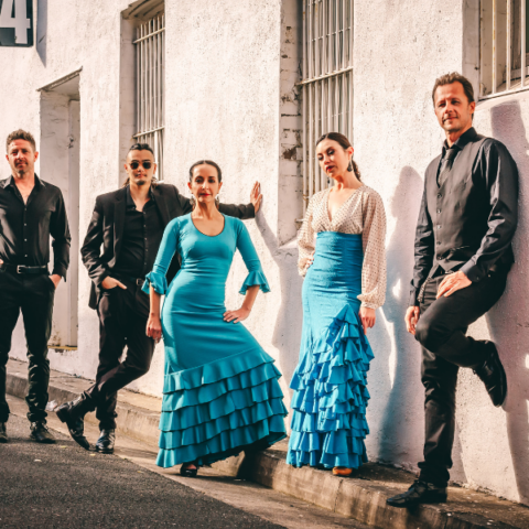 Flamenco group wearing traditional outfits resting upon an off white brick wall.