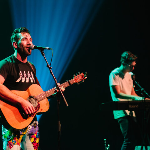 Aspy Jones is standing in front of a microphone on a stand holding his guitar. He is wearing a black t-shirt with colourful pants. There is a person standing behind Aspy wearing a white shirt playing the keyboard. 