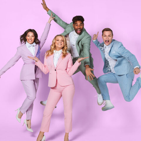 Two men and two women are dancing and leaping in a showbiz style, they are each wearing a neat suit in a pastel colour blue, pink, green and purple, there is a pink background. 