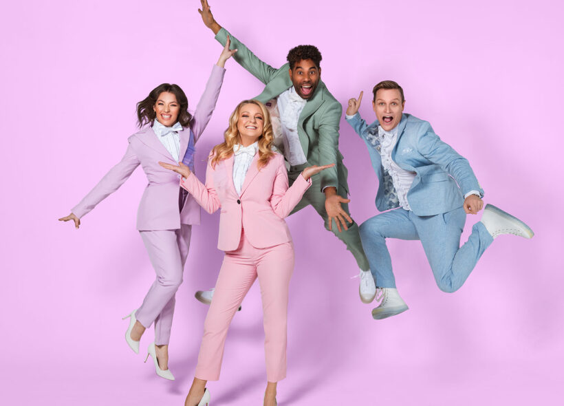 Two men and two women are dancing and leaping in a showbiz style, they are each wearing a neat suit in a pastel colour blue, pink, green and purple, there is a pink background. 