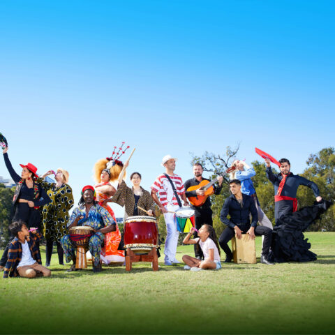 A group of people in brightly coloured costumes are dancing and holding a range of instruments. They are in a green grassy park with a bright blue sky.