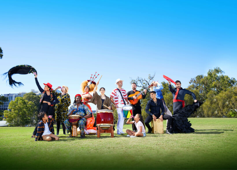 A group of people in brightly coloured costumes are dancing and holding a range of instruments. They are in a green grassy park with a bright blue sky.