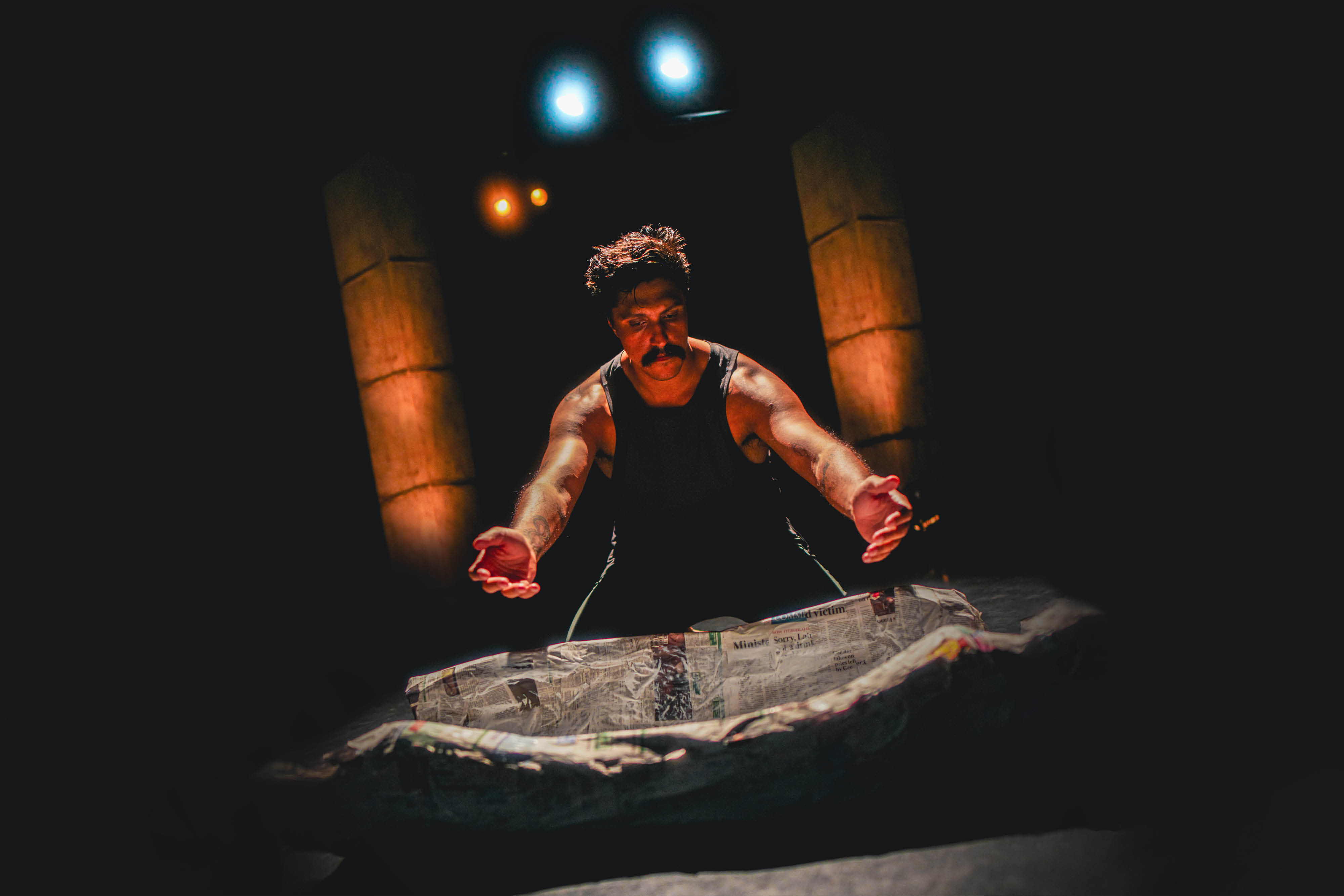 A man stands with arms outstretched above a coolomon vessel made from paper mache. He is spotlighted by stage lights and surrounded by darkness. 