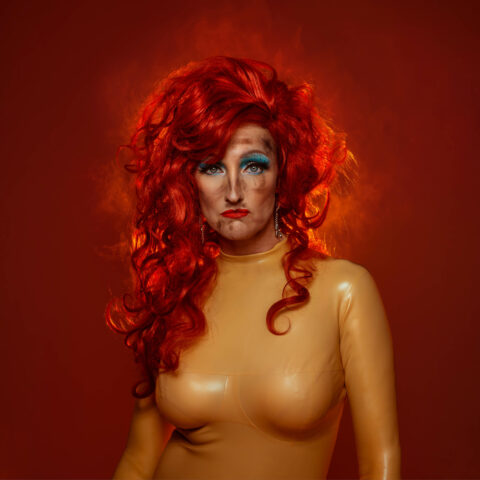 A woman glares at the camera, she has a huge amount of bright red hair, blue eyeshadow and is wearing a beige latex suit 