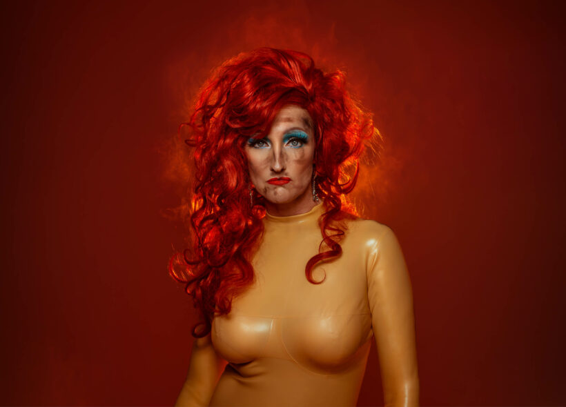 A woman glares at the camera, she has a huge amount of bright red hair, blue eyeshadow and is wearing a beige latex suit 
