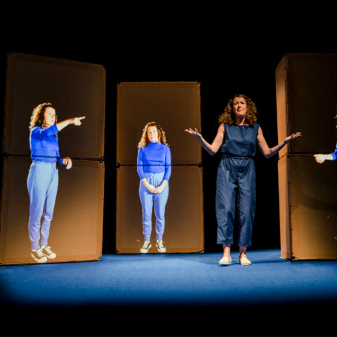 A woman stand son stage with her hands out in a 'why' posture, and three panels show projected images of her in different poses. 