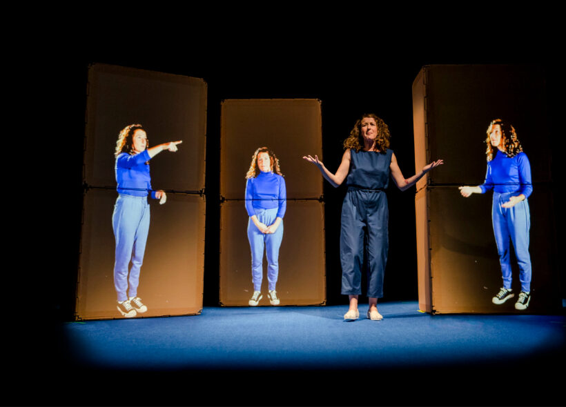A woman stand son stage with her hands out in a 'why' posture, and three panels show projected images of her in different poses. 