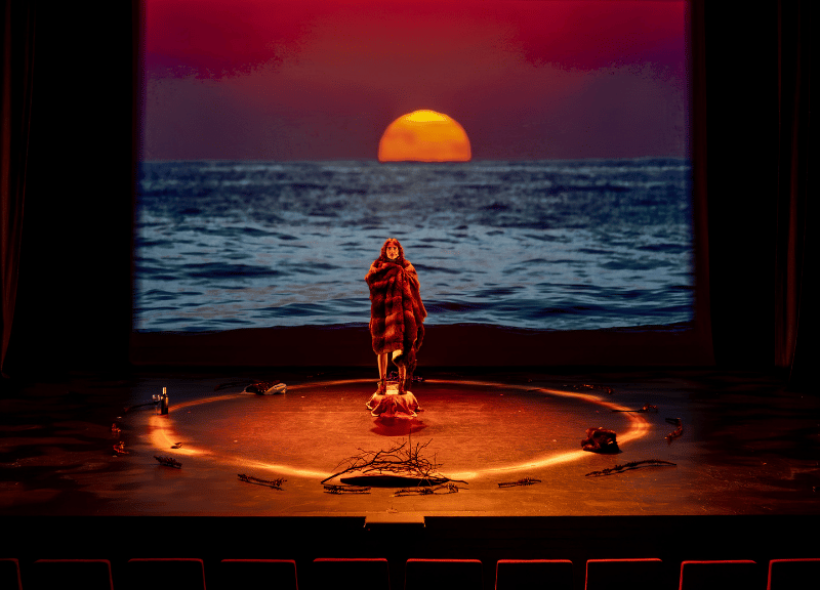 A figure draped in a fur coat stands in a spotlight within a circle of objects on a dark stage, with a large image of a sunset over the ocean projected behind them.