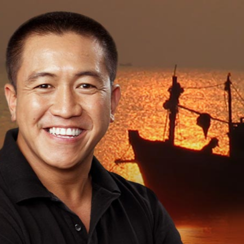 Smiling man in a black shirt stands in front of a scenic sunset background with a silhouette of a fishing boat on the water.