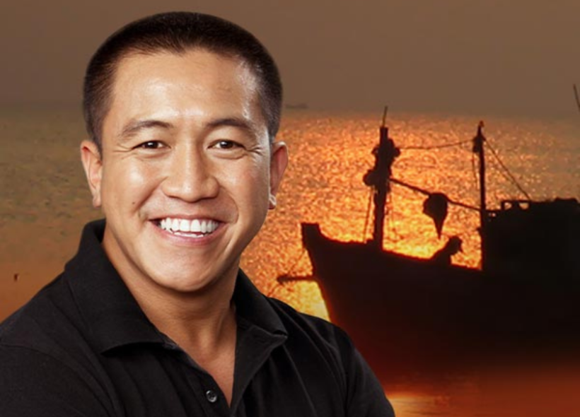 Smiling man in a black shirt stands in front of a scenic sunset background with a silhouette of a fishing boat on the water.