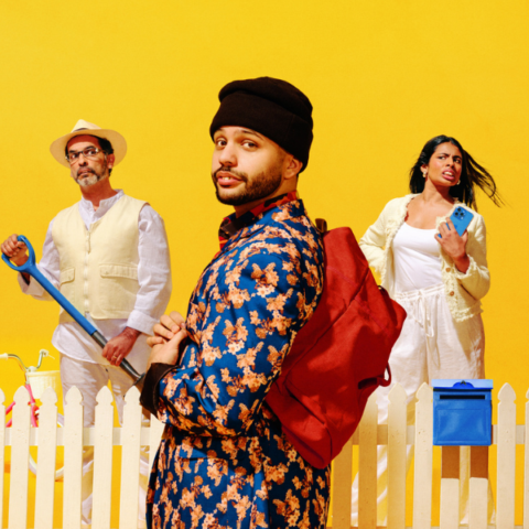 Three people stand in front of a white picket fence with a bright yellow background. One man wears floral print and a beanie, while the others wear light clothing and hold garden tools.
